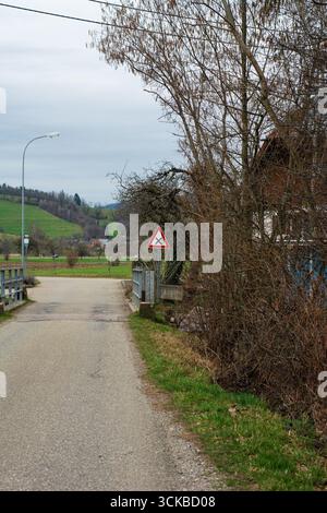 Eine einsame Straße führt zu einem Warnschild, flankiert von blattlosen Bäumen und grünen Hügeln im Hintergrund, die die Ruhe des ländlichen Li einfangen Stockfoto