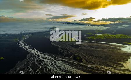 Aus der Vogelperspektive auf einen grünen kegelförmigen Hügel in Islands vulkanischem Gelände, mit weißen Flüssen, schwarzem Sand, einem Gletscher und schneebedeckten Bergen unter einer Führung Stockfoto