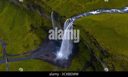 Blick aus der Vogelperspektive auf den Seljalandsfoss Wasserfall in Island, der von einer hohen Klippe in einen Pool darunter fällt, mit einem Wanderweg und üppigem Grün. Stockfoto