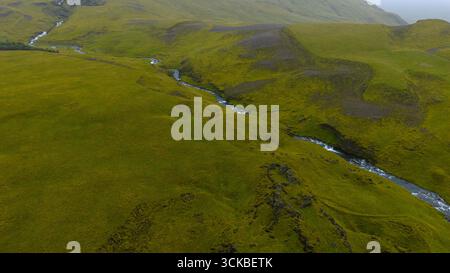 Aus der Vogelperspektive sehen Sie eine üppig grüne isländische Landschaft mit einem sich windenden Fluss, der durch moosbedeckte sanfte Hügel und unberührtes natürliches Gelände führt. Stockfoto