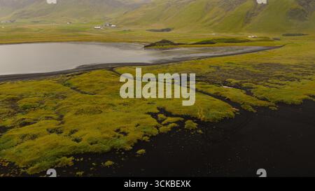 Luftbild eines reflektierenden Wasserkörpers, umgeben von schwarzem vulkanischem Sand, grünem Moos, sanften Hügeln, fernen Bergen und verstreuten Bauernhäusern. Stockfoto