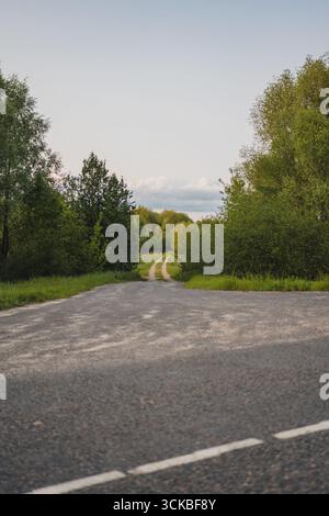 Eine ruhige ländliche Straße in Weißrussland übergeht von Pflaster zu Erde, eingerahmt von üppigen grünen Bäumen und Büschen unter einem klaren Himmel mit weichen Wolken. Stockfoto