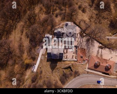 Ansammlung von Steinhäusern mit einzigartigen Dächern, umgeben von blattlosen Bäumen, einer gewundenen Straße mit einem blauen Auto und sichtbaren Ruinen auf unebenem Gelände. Stockfoto