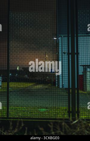 Moody-Nachtszene in Danzig mit einem hohen beleuchteten Industriebauwerk, einem Gitterzaun aus Metall und einer dezent beleuchteten urbanen Umgebung. Stockfoto
