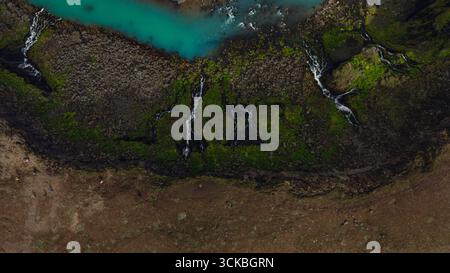 Der türkisfarbene Fluss fließt durch moosbedeckte vulkanische Felsen in Island, mit kaskadierenden Wasserfällen und einem kargen Erdgebiet, das darunter zu sehen ist. Stockfoto