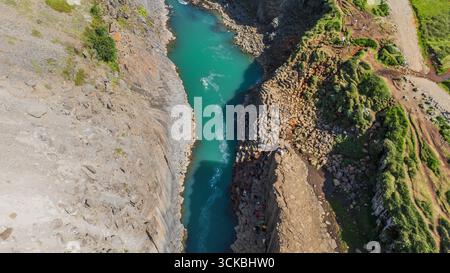 Türkisfarbener Fluss fließt durch einen tiefen Canyon mit Basaltfelswänden, rauem Gelände, grüner Vegetation und sichtbaren Wanderwegen in Island. Stockfoto