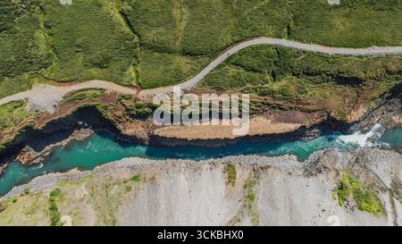 Ein sich windender türkisfarbener Fluss fließt durch zerklüftetes isländisches Gelände, das von felsigen Ufern, Grün und einer schmalen Straße umgeben ist. Stockfoto