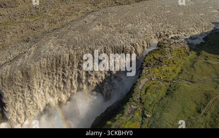 Der Dettifoss-Wasserfall stürzt kraftvoll aus der Luft, mit Nebel, einem schwachen Regenbogen, felsigem Gelände, grünem Moos, und ein gewundener Pfad sichtbar. Stockfoto