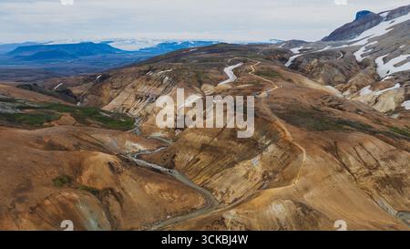 Blick aus der Vogelperspektive auf Islands Kerlingarfjoll-Gebirge mit Rhyolithhügeln, Schneefeldern, einer Feldstraße, einem Bach, und ferne Gletscher. Stockfoto