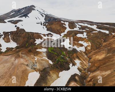 Aus der Vogelperspektive auf einen schneebedeckten Berg im isländischen Hochland, umgeben von mehrfarbigen Rhyolithhügeln, grünem Moos und kontrastierenden Erdtönen. Stockfoto
