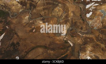 Aus der Vogelperspektive auf Islands karges Gelände mit gewundenen Flüssen, goldbraunen Hügeln, Ockertönen und Schneestücken in höheren Lagen. Stockfoto