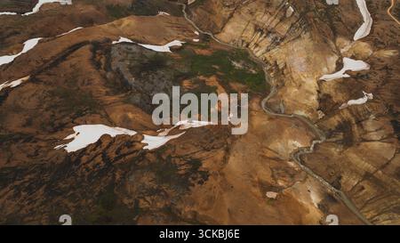 Ein dramatischer Blick aus der Luft auf Islands Hochland mit farbenfrohem geothermischem Gelände, verstreuten Schneefeldern und einem sich windenden Fluss, der durch die s schneidet Stockfoto