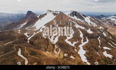 Dramatische Aussicht auf die zerklüfteten isländischen Berge mit schneebedeckten Streifen, rötlich braunem vulkanischem Gelände und einer gewundenen Feldstraße im Vorland Stockfoto
