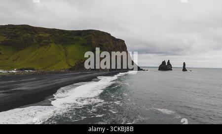 Reynisfjara Beach in Island bietet Basaltklippen, moosbedeckte Hänge und Reynisdrangar Meer stapelt sich unter einem bewölkten Himmel mit sanften Wellen. Stockfoto