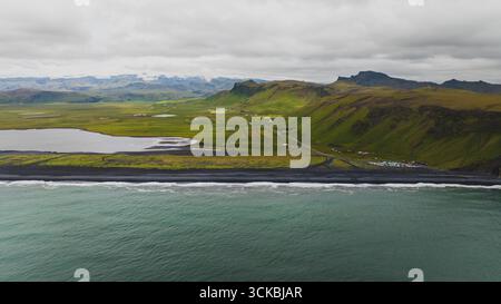 Dramatische Aussicht auf Islands Südküste mit schwarzem Sandstrand, grünen Feldern, zerklüfteten Hügeln, einer kleinen Siedlung und ruhigem Binnenwasser. Stockfoto