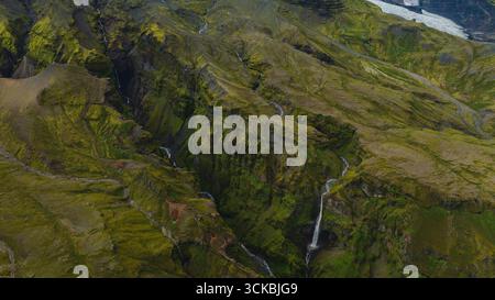 Dramatischer Blick aus der Luft auf einen moosbedeckten Canyon in Island mit kaskadierenden Wasserfällen, zerklüfteten Klippen und einem Gletscher, der im rechten oberen Maisbereich sichtbar ist Stockfoto