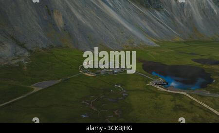 Eine kleine Siedlung befindet sich am Fuße eines steilen Berges in Island, umgeben von grünen Feldern, mit einer gewundenen Straße, die zu einem lebhaften blauen See führt. Stockfoto