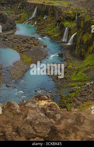 Ein sich windender türkisfarbener Fluss fließt durch eine zerklüftete isländische Schlucht, mit kleinen Wasserfällen, die moosbedeckte Klippen hinabstürzen und vulkanisches Felsformat Stockfoto