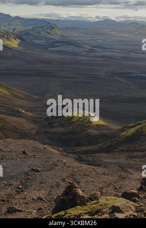 Ein riesiges vulkanisches Gelände in Island mit dunkler Erde, grünen Moosflecken, sanften Hügeln, Tälern und fernen Bergen unter einem bewölkten Himmel. Stockfoto
