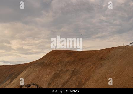 Eine Person steht auf einem rostfarbenen Kamm unter einem bewölkten Himmel, umgeben von kargen, erodierten Hängen in einer typisch für Island Vulkanlandschaft. Stockfoto