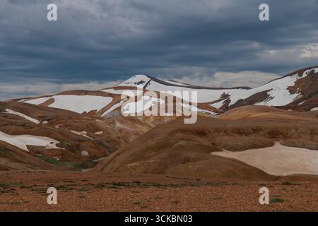 Leuchtende rote, orange und braune Rhyolithberge in Landmannalaugar, Island, mit Schneeflächen unter bedecktem Himmel im isländischen Hochland. Stockfoto