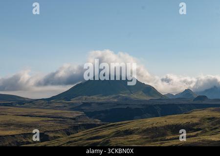 Ein Berg in Island, der teilweise von niedrigen Wolken bedeckt ist, umgeben von grünen moosigen Ebenen, dunklem vulkanischem Gelände und Canyons unter einem klaren blauen Himmel. Stockfoto