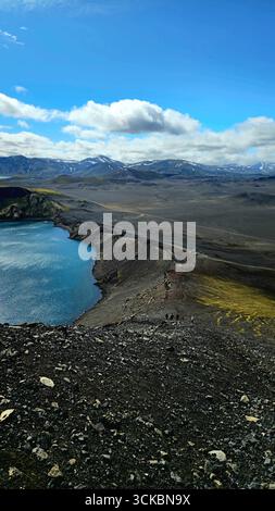 Ein lebhafter blauer vulkanischer Kratersee in Island, umgeben von dunklem felsigem Gelände. Schneebedeckte Berge und kleine Figuren verleihen der Szene einen besonderen Akzent. Stockfoto