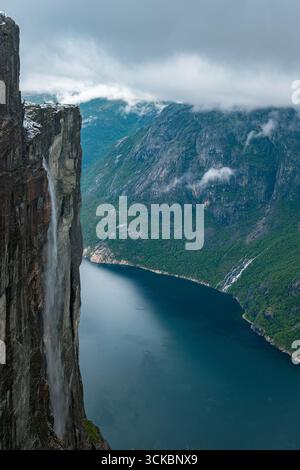 Dramatische Klippe mit Blick auf Lysefjord in Norwegen, mit einem Wasserfall, zerklüfteten bewaldeten Bergen, Nebel und tiefblauem Wasser unter einem bewölkten Himmel Stockfoto