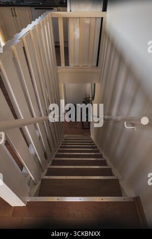 Eine schmale Treppe mit Holztreppen und weißen Geländern führt zu einer Landung mit schwarzem Rucksack und Ablage. Oben ist eine Küchenecke zu sehen. Stockfoto