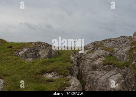 Ein kleiner weißer Leuchtturm mit rotem Dach steht auf zerklüftetem felsigem Gelände mit grünen Grasflecken unter einem bewölkten Himmel auf der Insel Utsira, NOR Stockfoto