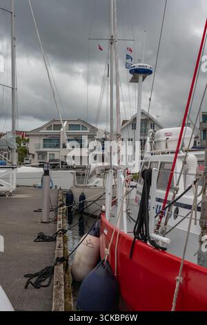 Segelboote legten an einem Betonpier in Norwegen an, mit einem roten Boot im Vordergrund. Weiße Gebäude mit großen Fenstern säumen das bedeckte Wasser Stockfoto