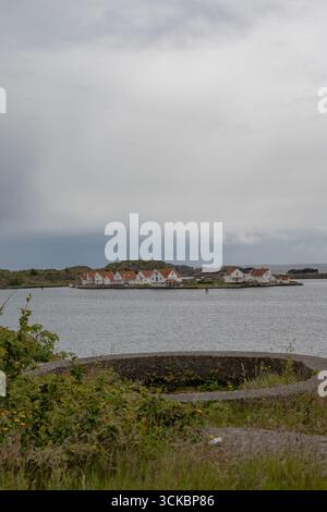 Eine felsige Insel in Norwegen mit weißen Häusern und roten Dächern, umgeben von ruhigem Wasser. Eine grasbewachsene Fläche mit einer kreisförmigen Betonkonstruktion steht im Vordergrund Stockfoto
