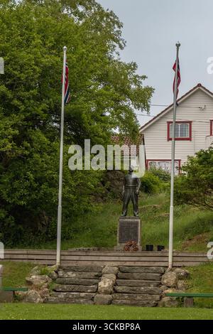 Eine Statue auf einem Steinsockel mit norwegischen Fahnen, umgeben von Grün und Bäumen. Ein weißes Haus mit roter Zierleiste und einem gekachelten Dach ist in der Ba zu sehen Stockfoto