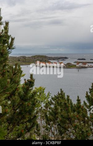 Eine Reihe weißer Häuser mit roten Dächern befindet sich an einem schmalen Küstenstreifen in Norwegen, eingerahmt von Kiefern, felsigem Gelände und einem bewölkten Himmel. Stockfoto