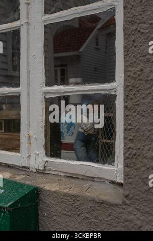 Nahaufnahme eines verwitterten, weiß gerahmten Fensters mit grünem Briefkasten, das Häuser im norwegischen Stil in einer Küstenstadt widerspiegelt. Stockfoto