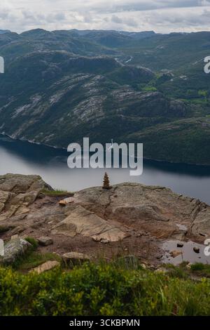 Blick auf Lysefjord in Norwegen mit einem Steinhaufen auf einem Felsvorsprung, einem entfernten Wasserfall, zerklüfteten Bergen und ruhigem, reflektierendem Wasser unter einem bewölkten Himmel Stockfoto