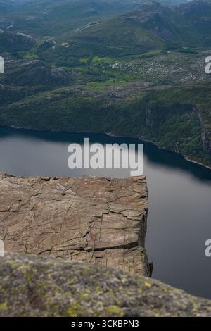 Blick auf Preikestolen's zerklüftete flache Spitze, mit dem ruhigen Wasser des Lysefjords und grünen Hügeln im Hintergrund, mit kleinen Siedlungen und natürlichen Scener Stockfoto