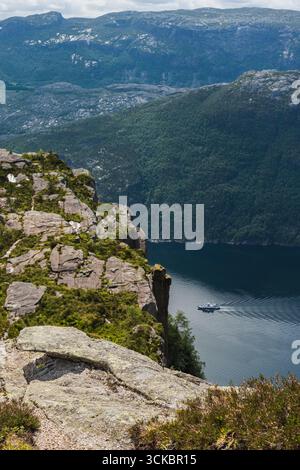 Blick auf Preikestolen in Norwegen, mit zerklüfteten Klippen, einem ruhigen Fjord mit einem fahrenden Boot und Bergen mit felsigem und bewaldetem Gelände. Stockfoto