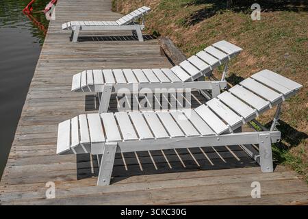 Ein rustikales hölzernes Dock mit einem weißen Loungesessel und einem Beistelltisch sitzt friedlich an einem ruhigen See Stockfoto