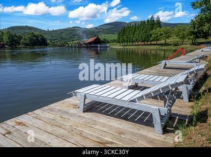 Ein rustikales hölzernes Dock mit einem weißen Loungesessel und einem Beistelltisch sitzt friedlich an einem ruhigen See Stockfoto