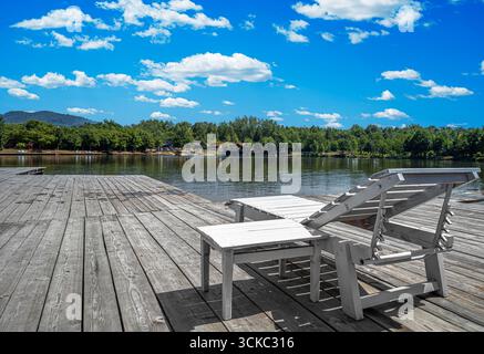 Ein rustikales hölzernes Dock mit einem weißen Loungesessel und einem Beistelltisch sitzt friedlich an einem ruhigen See Stockfoto