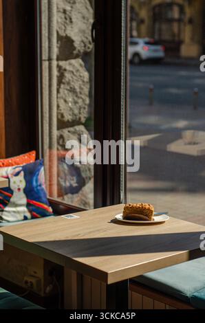 Ein Stück Karottenkuchen auf einem hölzernen Café-Tisch am Fenster mit bunten dekorativen Kissen und Sonnenlicht, gemütliches, urbanes Café-Interieur. Stockfoto