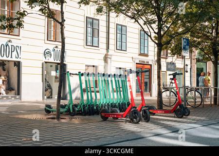 Ljubljana Slowenien - 23. August 2025: Reihe geparkter Elektroroller auf der Stadtstraße mit Bäumen und Gebäuden im Hintergrund. Stockfoto