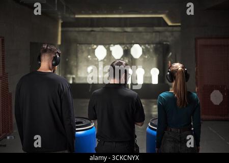 Drei junge Erwachsene, zwei kaukasische Männer und eine kaukasische Frau, die Seite an Seite stehen und schützende Ohrenschützer tragen, bereiten sich darauf vor, auf Papierziele im Schießstand zu schießen Stockfoto