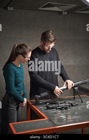 Kaukasischer junger erwachsener Mann demonstrierte der jungen kaukasischen erwachsenen Frau, die beide eine Schutzbrille trugen und mit Schusswaffenteilen im Innenbereich am Tisch standen Stockfoto