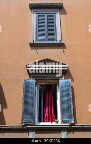 Orangefarbene Wand mit blauen Fensterläden und roten Vorhängen mit zwei Fenstern und einer Pflanze Stockfoto