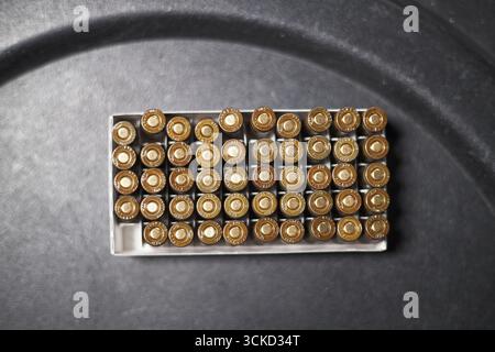 Schachtel mit Messing-Handfeuerpatronen, die in ordentlichen Reihen auf dunkler Oberfläche angeordnet sind, mit Metallgehäusen und Grundierungen von oben, die Munitionslager und Zubehör für Schusswaffen zeigen Stockfoto