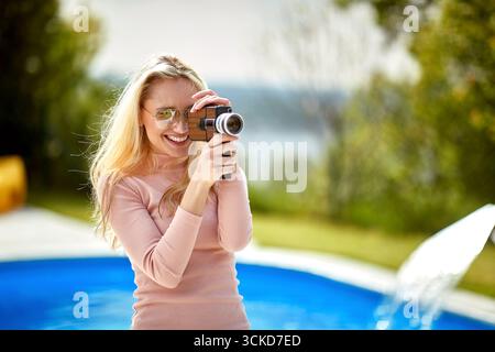 Junge, fröhliche blonde Frau, die eine Videokamera hält und draußen am Pool in der Natur aufnimmt, fühlt sich positiv. Lifestyle, Spaß, Aktivitätskonzept. Stockfoto