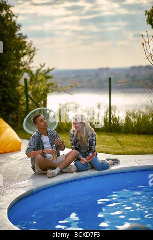 Junges Paar, das draußen am Pool sitzt und ein fröhliches Gespräch führt und den Sonnenuntergang im Sommer genießt. Zweisamkeit, Spaß, Liebeskonzept. Stockfoto