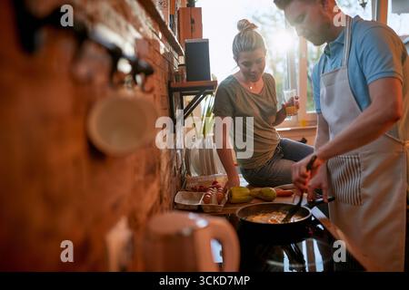 Der Mann macht Frühstück in einer Pfanne, Rührei, während die Frau auf der Arbeitsplatte sitzt und ansieht, was er macht. Zweisamkeit, Lebensstil, häuslich Stockfoto